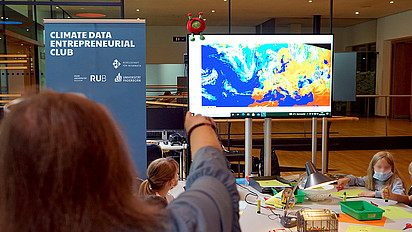 Jugendliche bei einem interaktiven Workshop. Auf dem Bildschirm in der Mitte des Raumes sind Satellitendaten angezeigt.